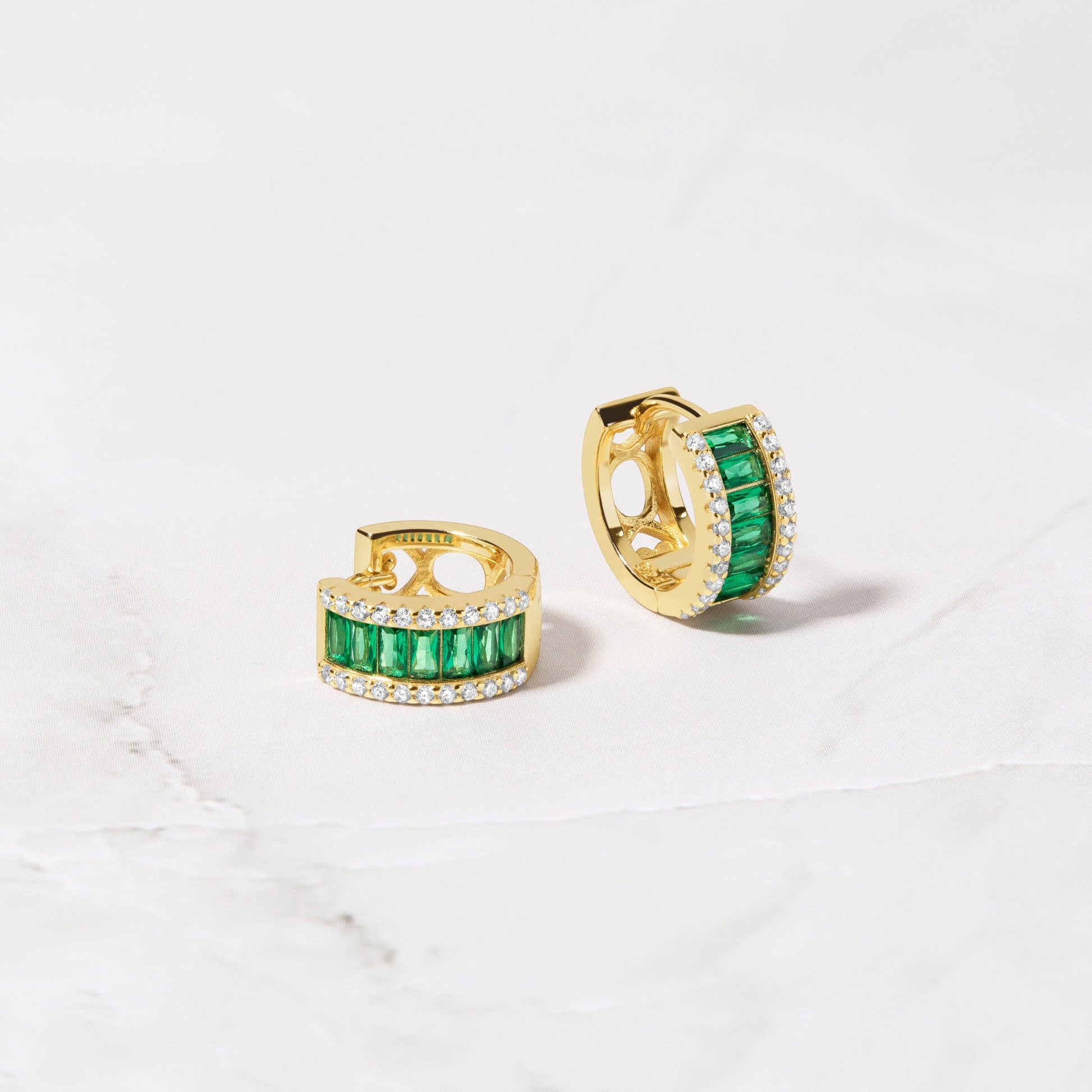 A pair of small gold-plated sterling silver huggie hoops witting on a white stone surface. Each hugggie is set with a column of horizontal baguette cut green cubic zirconia. On either side of this column are small white cubic zirconia. One huggie is facing slightly to the right showing a circular gold detail on the back of the earring. The other earring is sitting on its side.