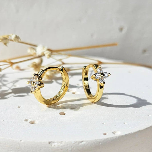 A pair of small gold-plate sterling silver huggie hoop earrings sitting on a white stone surface. The earrings have a small flower detail. The flower is made of 4 white cubic zirconia with surrounding a gold center. 