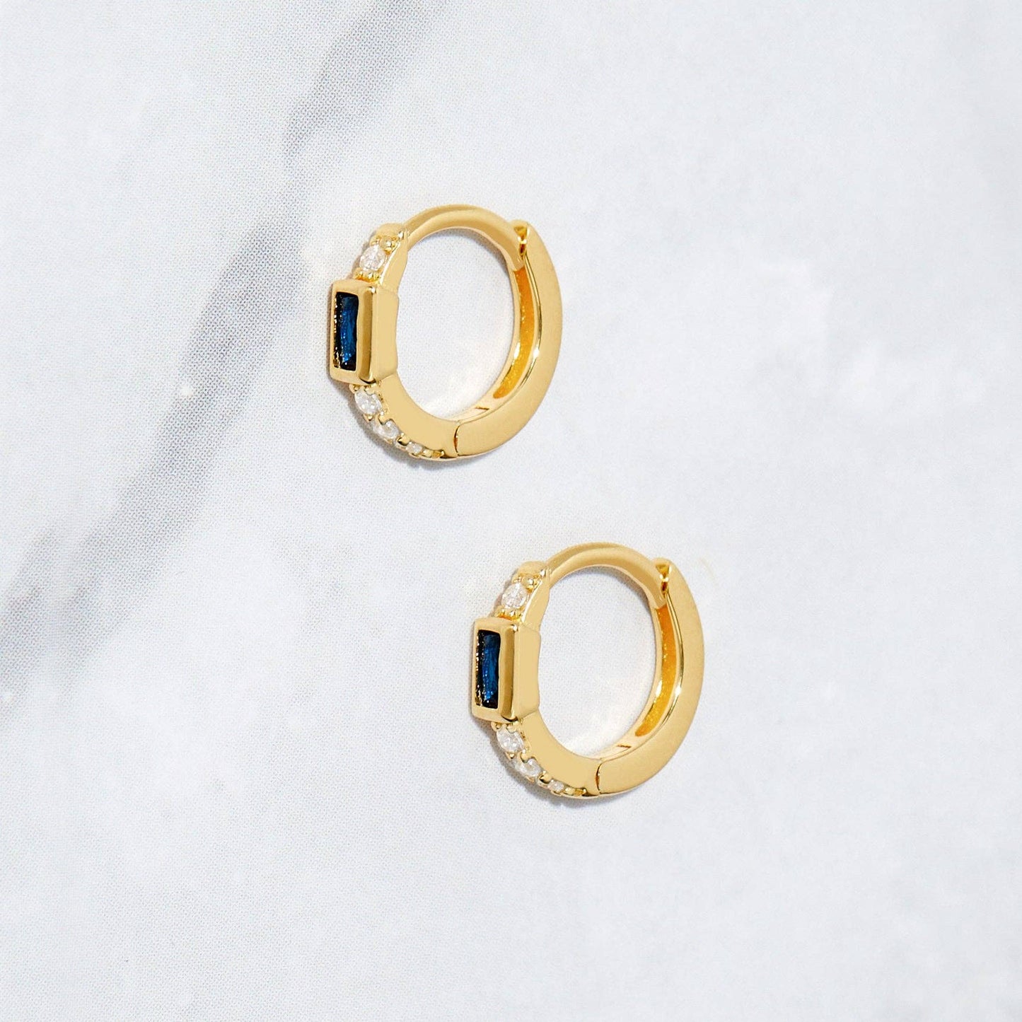 Side profile of a pair of small gold-plated huggie hoop earrings with a blue baguette-shaped cubic circonia. There is one white cubic zirconia above the blue stone and a line of 3 white cubic zirconias below. 