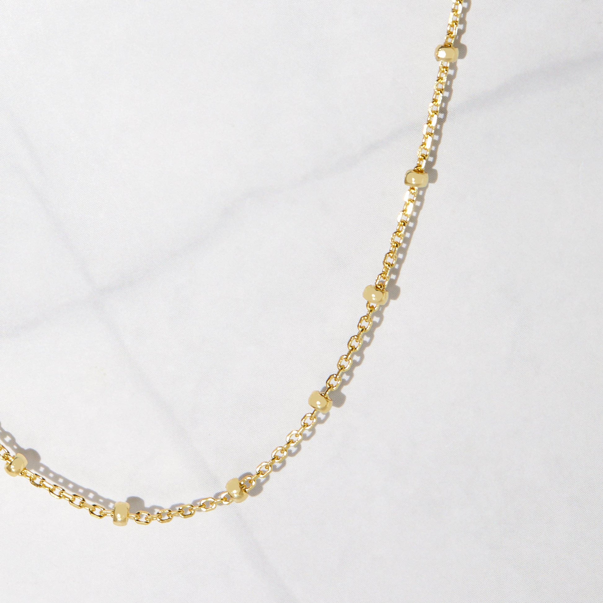Close up of a dainty gold-plated sterling silver chain with gold beads hanging against a white stone background. The beads are separated by one inch of chain. 