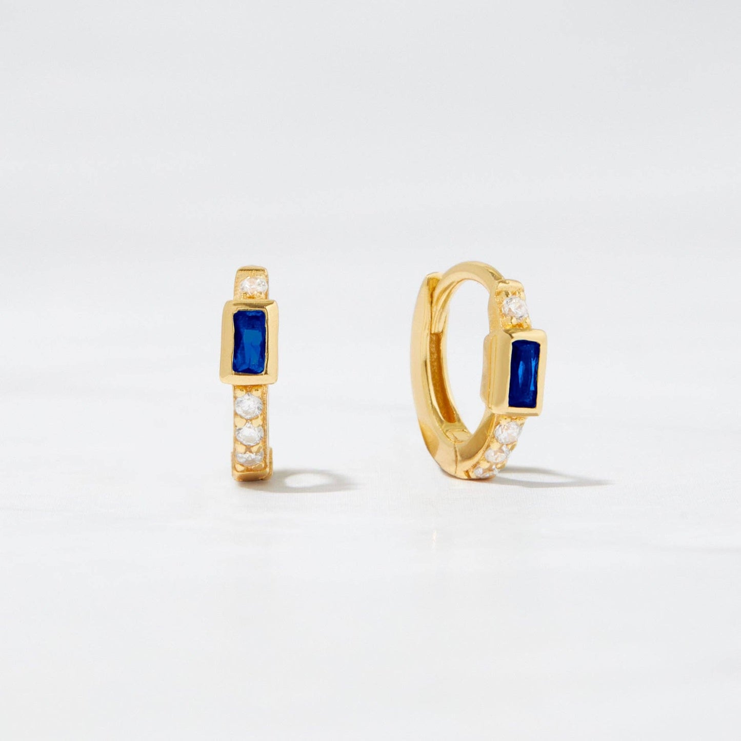 Small gold-plated huggie hoop earrings with a blue baguette-shaped cubic circonia. There is one white cubic zirconia above the blue stone and a line of 3 white cubic zirconias below. 