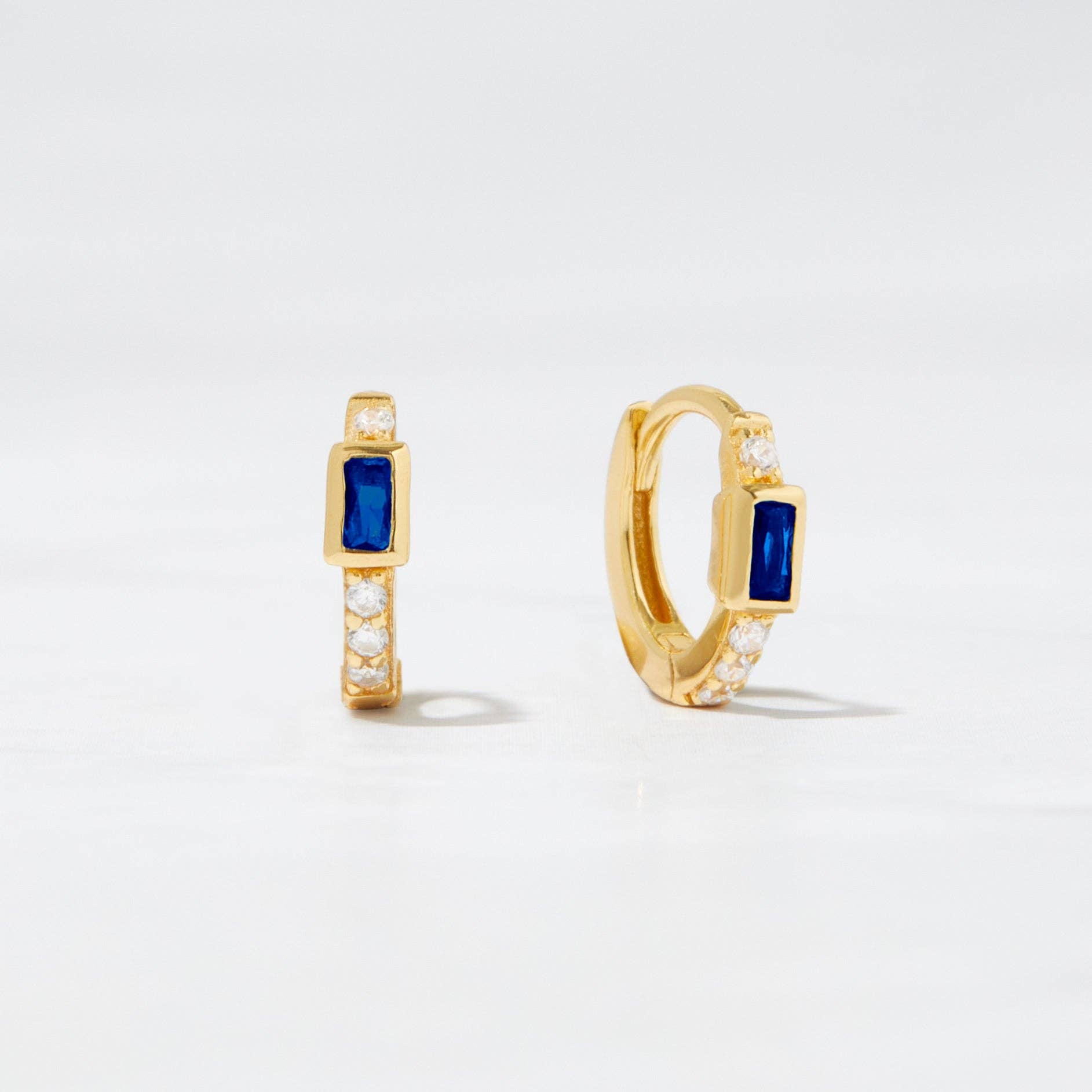 Small gold-plated huggie hoop earrings with a blue baguette-shaped cubic circonia. There is one white cubic zirconia above the blue stone and a line of 3 white cubic zirconias below. 